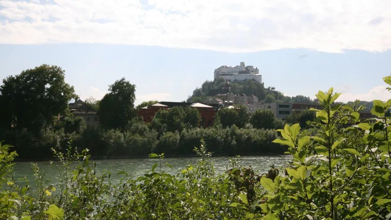 Panoramablick auf die Festung Hohensalzburg über dem Fluss, umgeben von üppiger Natur.