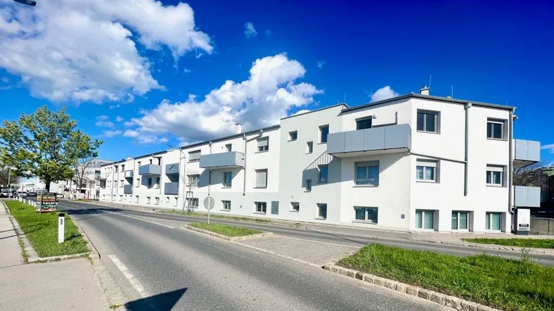 Moderne Wohnanlage mit weißer Fassade und grauen Balkonen entlang einer Straße unter blauem Himmel.