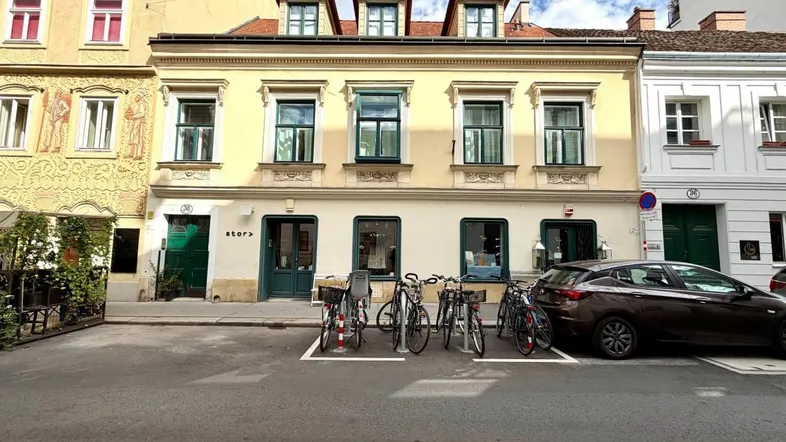 Ansicht der Gebäudefassade mit Geschäften im Erdgeschoss, Fahrrädern und einem geparkten Auto auf der Straße.