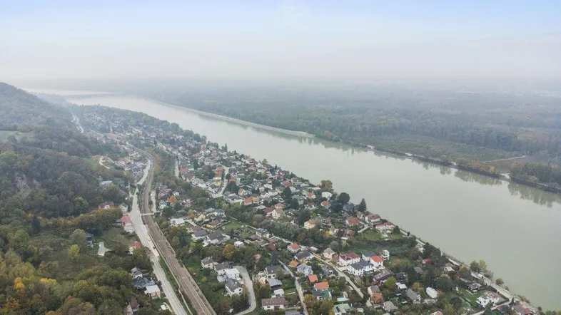 Luftaufnahme einer Siedlung entlang eines Flusses mit bewaldeten Hügeln im Hintergrund.