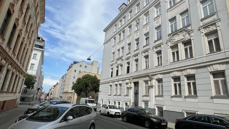 Straße mit parkenden Autos und Gebäuden im Wiener Stil unter blauem Himmel.
