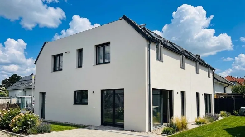 Modernes Einfamilienhaus mit weißer Fassade, dunklem Dach und gepflegtem Vorgarten unter blauem Himmel.