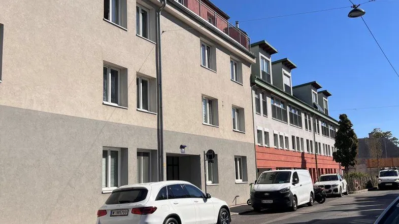 Mehrstöckiges Wohngebäude mit heller Fassade und geparkten Autos an einer sonnigen Straße.