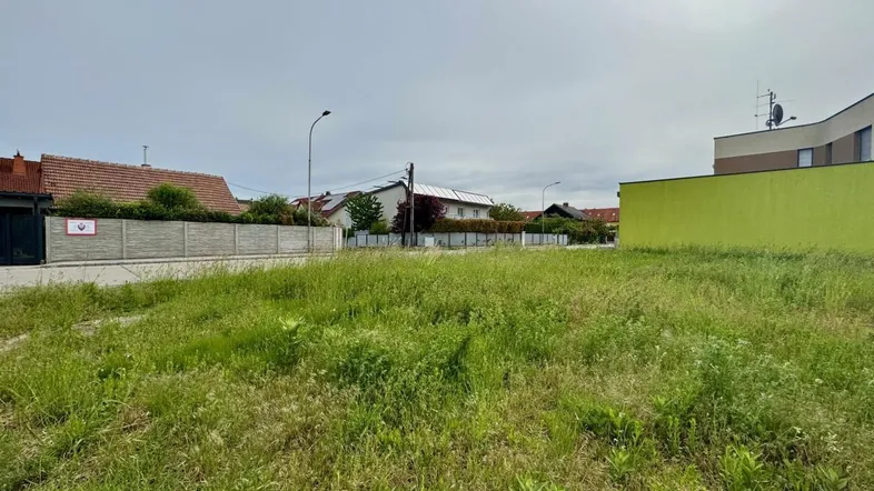 Unbebautes Grundstück mit hohem Gras, umgeben von Zäunen und Wohnhäusern unter bewölktem Himmel.