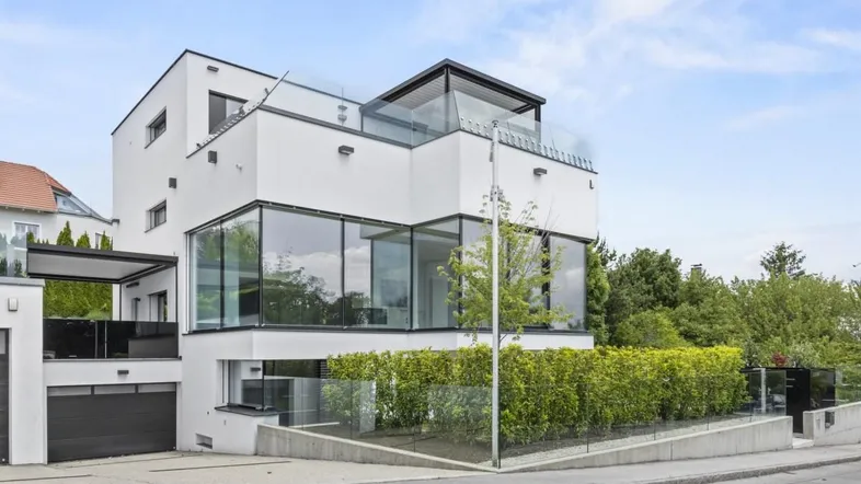 Moderne Villa mit weißer Fassade, großen Glasflächen und gepflegter Hecke an der Straße.