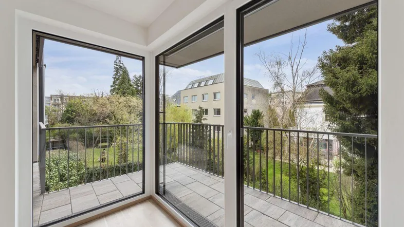 Heller Balkon mit Fliesenboden und Blick auf den grünen Innenhof und umliegende Gebäude.