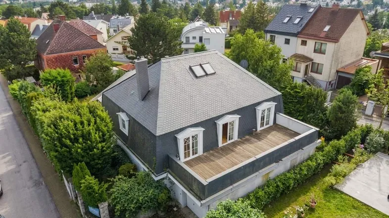 Luftaufnahme eines Hauses mit dunklem Schieferdach und großer Holzterrasse in grüner Umgebung.