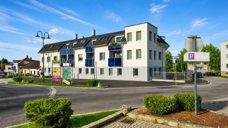 Modernes Mehrfamilienhaus an einer Kreuzung mit blauen Balkonen und gepflegter Umgebung.