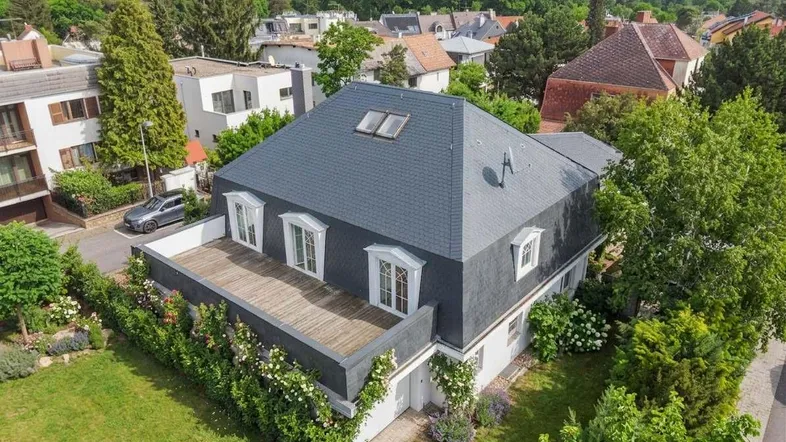 Luftaufnahme einer eleganten Villa mit Mansarddach und großer Holzterrasse, umgeben von viel Grün.