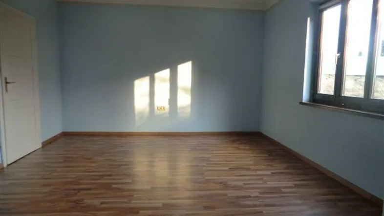 Helles Zimmer mit blauen Wänden und Laminatboden, durch ein Fenster fällt Sonnenlicht herein.
