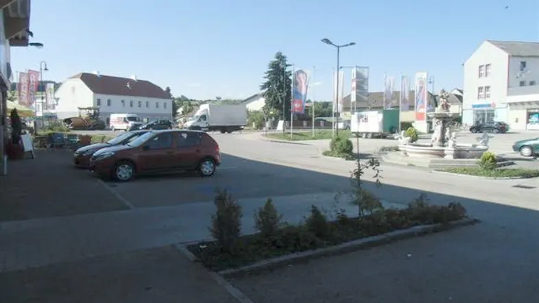 Blick auf eine Dorfstraße mit parkenden Autos, Gebäuden und einem Brunnen im Zentrum.