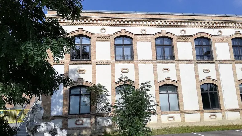Historische Fassade mit markanten Rundbogenfenstern und dekorativen Elementen, umgeben von Grünflächen.