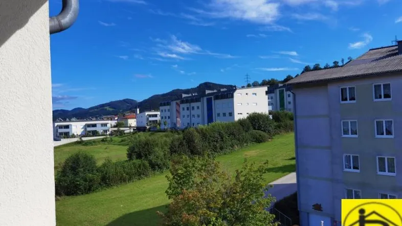 Blick von einem Balkon auf Wohngebäude und grüne Hügel unter blauem Himmel.