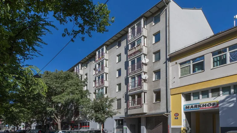 Mehrstöckiges Wohngebäude mit Balkonen und Geschäften im Erdgeschoss an einer städtischen Straße.