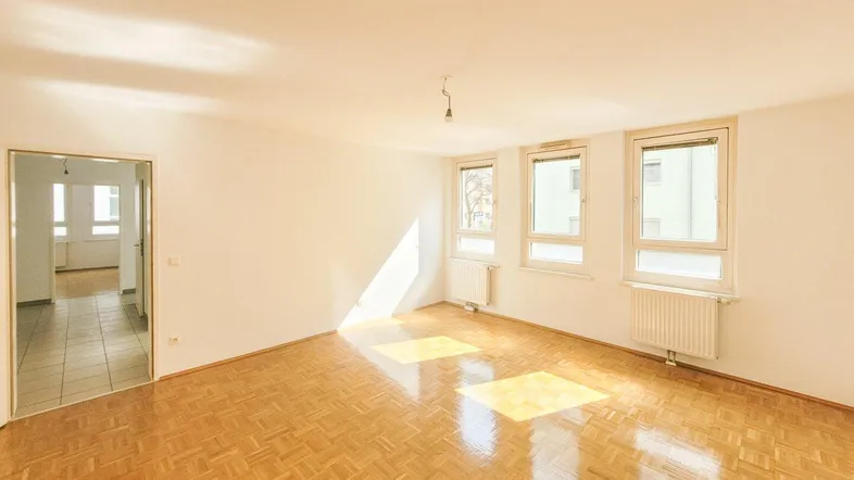 Heller, leerer Raum mit Fischgrätparkettboden und großen Fenstern, durch die Sonnenlicht einfällt.