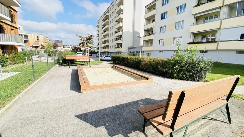 Gemeinschaftlicher Außenbereich mit Sandkasten und Sitzbänken, umgeben von Wohngebäuden unter blauem Himmel.