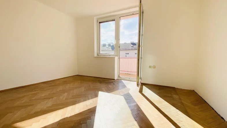 Heller Raum mit Fischgrätparkett und Zugang zu einem Balkon, durchflutet von natürlichem Sonnenlicht.