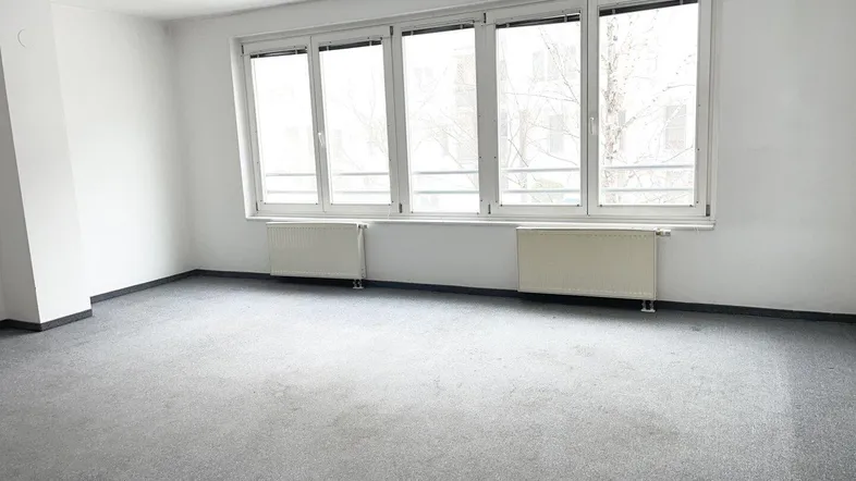 Leeres Wohnzimmer mit grauem Teppichboden, weißen Wänden und großen Fenstern, die viel Licht hereinlassen.