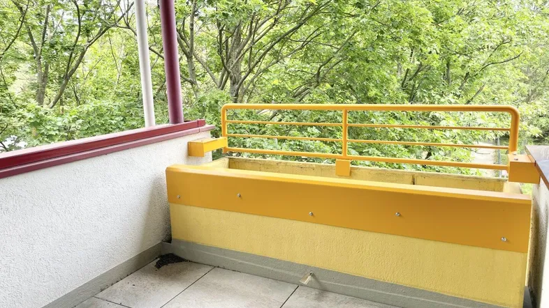Balkon mit gelbem Geländer und Fliesenboden, umgeben von üppigem Grün.