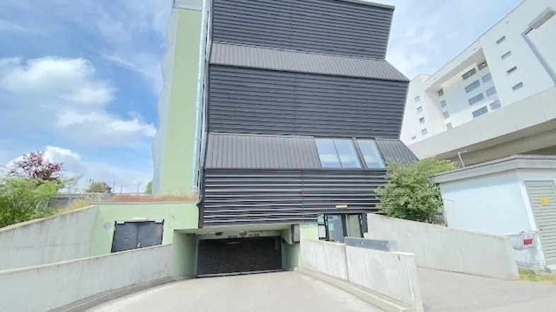 Modernes Gebäude mit dunkler Fassade und Zufahrt zur Tiefgarage unter blauem Himmel.