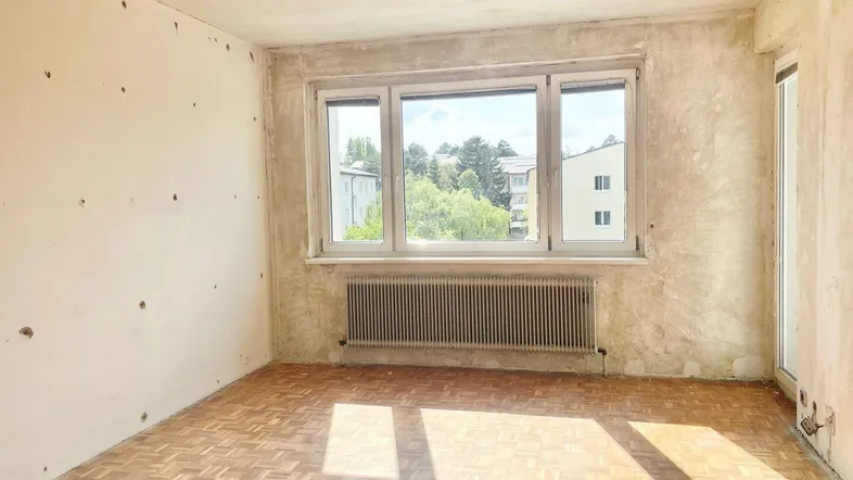Unfertiges Zimmer mit Parkettboden, Heizkörper und Blick ins Grüne, bereit zur Renovierung.