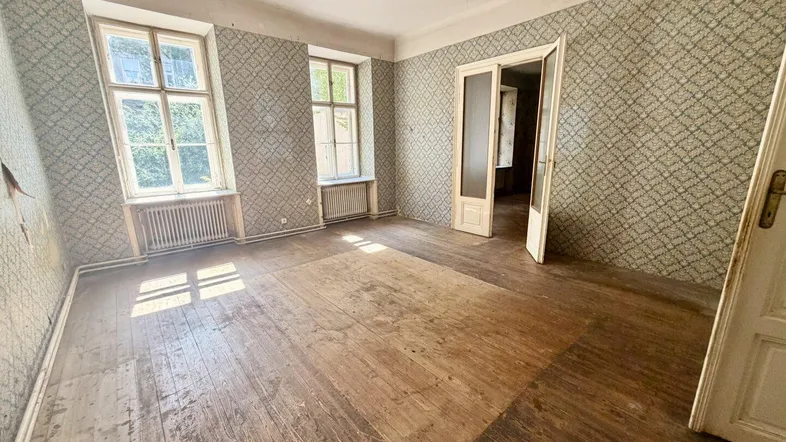Geräumiges Zimmer mit abgenutztem Holzboden und gemusterter Tapete, bereit für eine Renovierung.
