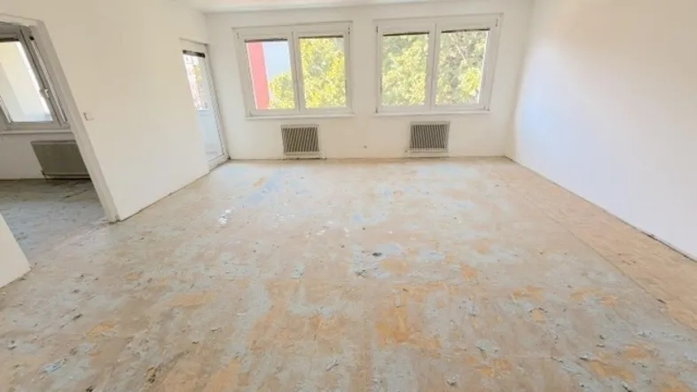 Helles Zimmer mit weiß gestrichenen Wänden, altem Bodenbelag und Zugang zu einem Balkon.