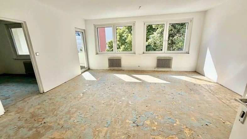 Heller Raum mit renovierungsbedürftigem Boden, Blick ins Grüne und Balkonzugang.