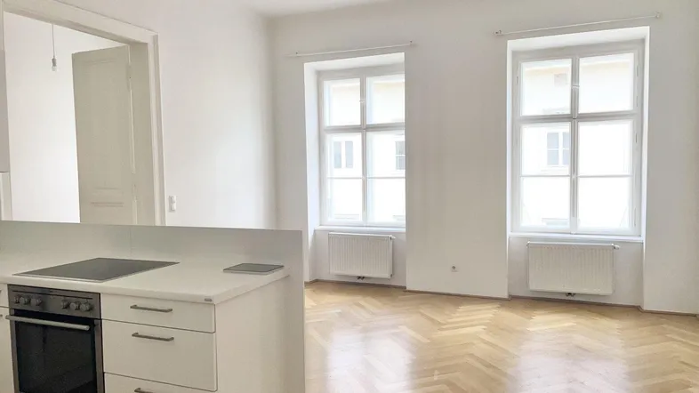 Heller Wohnbereich mit Fischgrätparkett, zwei großen Fenstern und integrierter Küchenzeile.