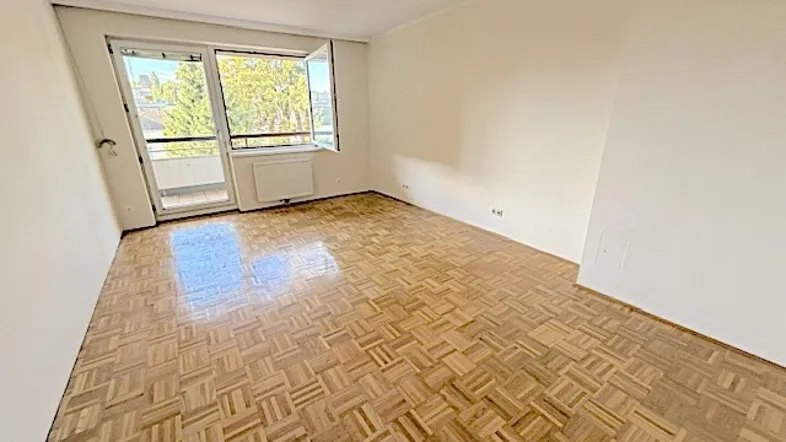 Helles, leeres Zimmer mit Fischgrätparkett und Zugang zu einem Balkon.