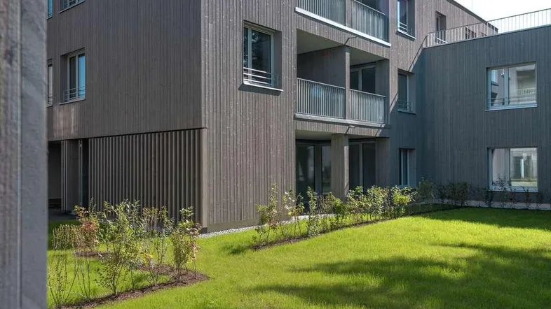 Modernes Gebäude mit Holzfassade, Balkonen und gepflegtem Rasen im Vordergrund unter blauem Himmel.
