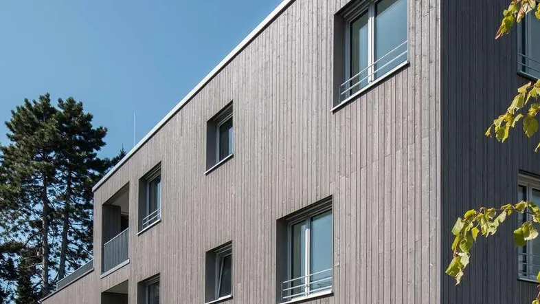 Moderne Wohnanlage mit Holzfassade und großen Fenstern unter blauem Himmel.