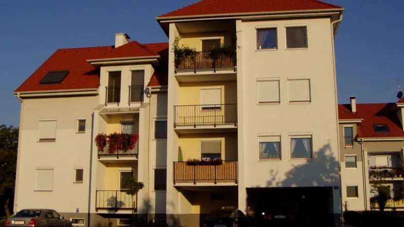 Mehrfamilienhaus mit rotem Ziegeldach, hellen Fassaden und mehreren Balkonen mit Geländern.
