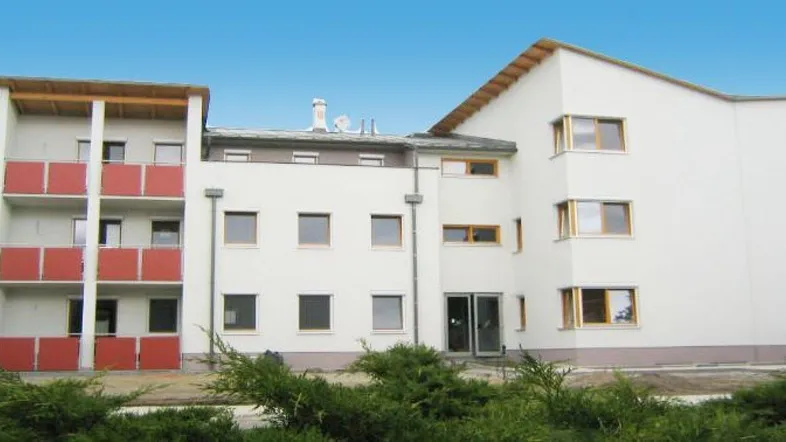 Modernes Mehrfamilienhaus mit weißer Fassade, roten Balkonen und grüner Bepflanzung im Vordergrund.