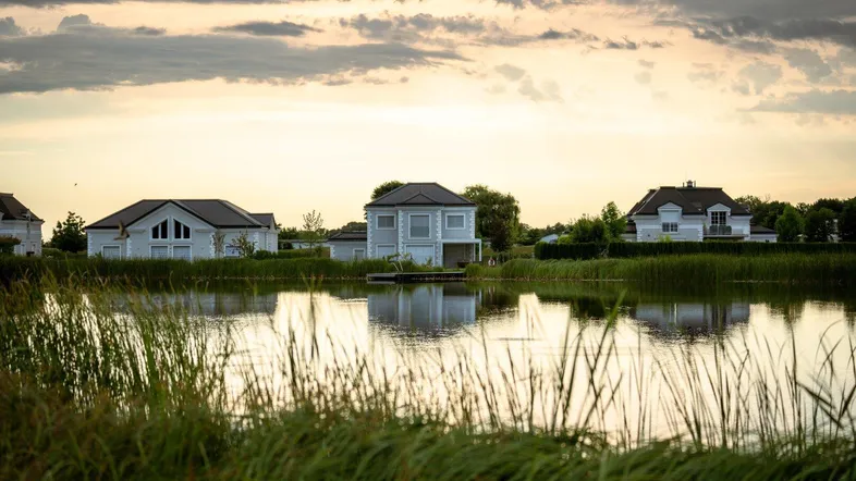 Häuser am Seeufer mit Schilf im Vordergrund bei Sonnenuntergang und ruhiger Wasseroberfläche.