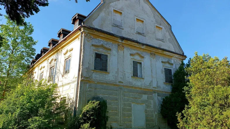 Altes, verputztes Gebäude mit mehreren Fenstern und Mansarddach in ländlicher Umgebung.