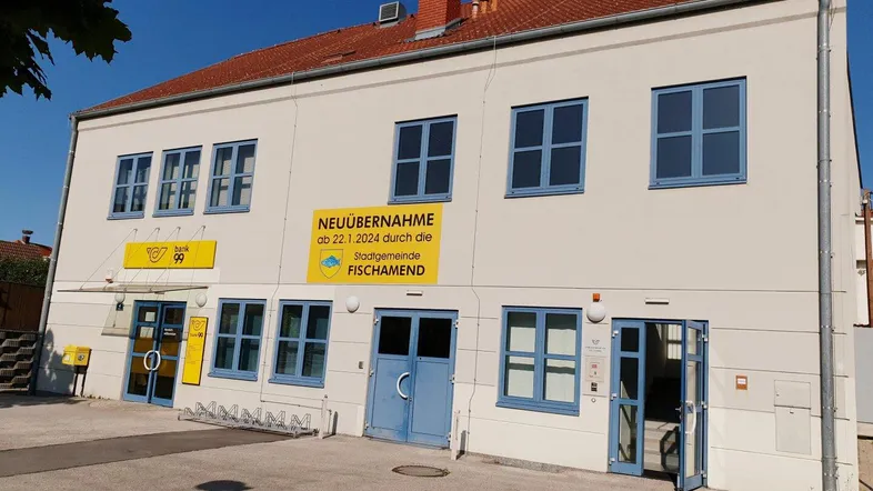 Gebäudeaußenansicht mit heller Fassade, blauen Fenstern und einem Schild zur Neuübernahme durch die Stadtgemeinde.