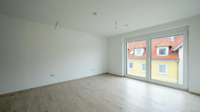Helles, leeres Zimmer mit großen Fenstern, hellem Holzboden und Blick auf ein Nachbargebäude.