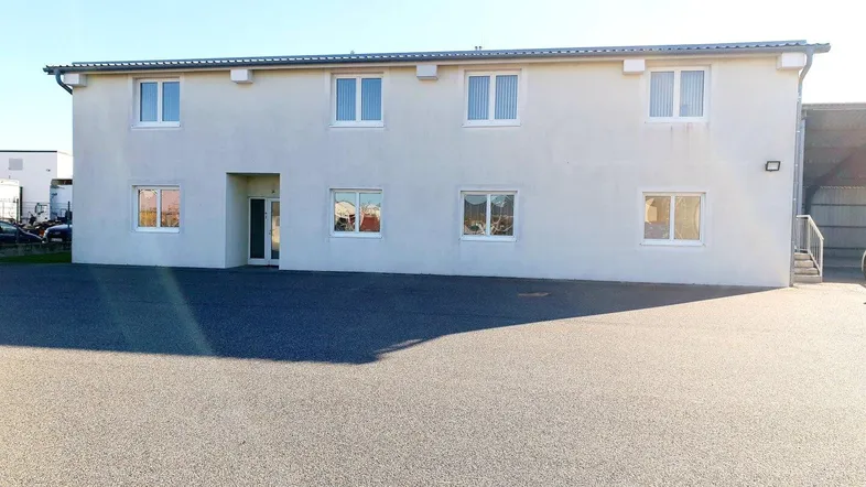 Vorderansicht des Gebäudes mit mehreren Fenstern und einem großen asphaltierten Parkplatz davor.