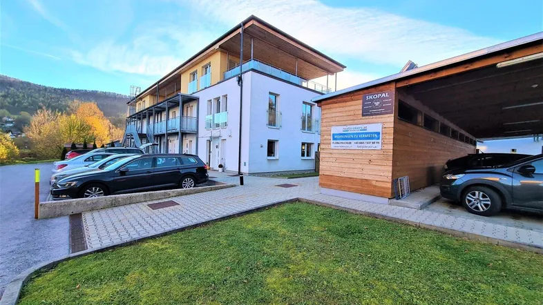 Modernes Mehrfamilienhaus mit Balkonen und überdachten Parkplätzen in bergiger Umgebung.