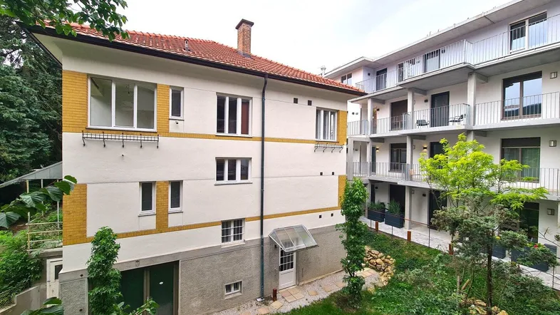 Ansicht von zwei Gebäuden, einem älteren mit gelben Akzenten und einem modernen Neubau mit Balkonen, umgeben von viel Grün.
