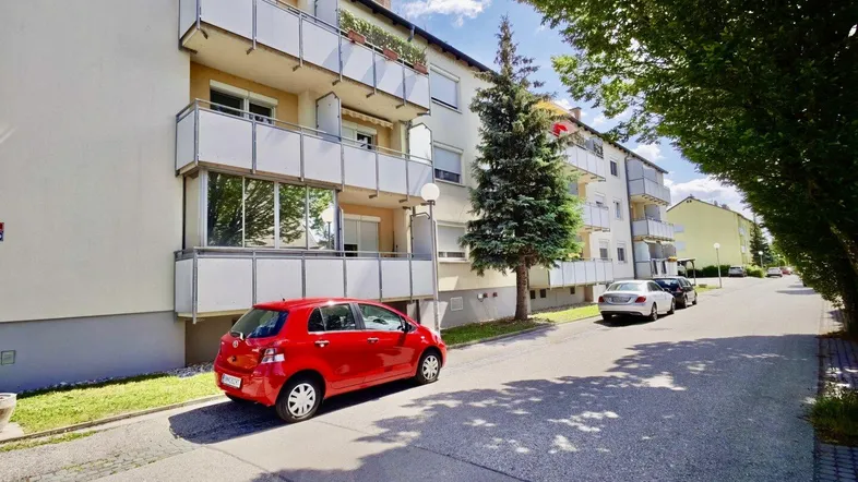 Mehrfamilienhaus mit mehreren Balkonen und parkenden Autos auf der Straße unter blauem Himmel.
