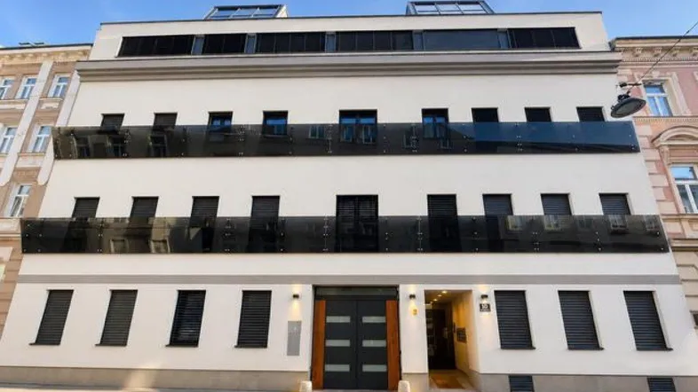 Moderne Gebäudefassade mit großen Fenstern, schwarzen Rollläden und Glasbalkonen in städtischer Umgebung.