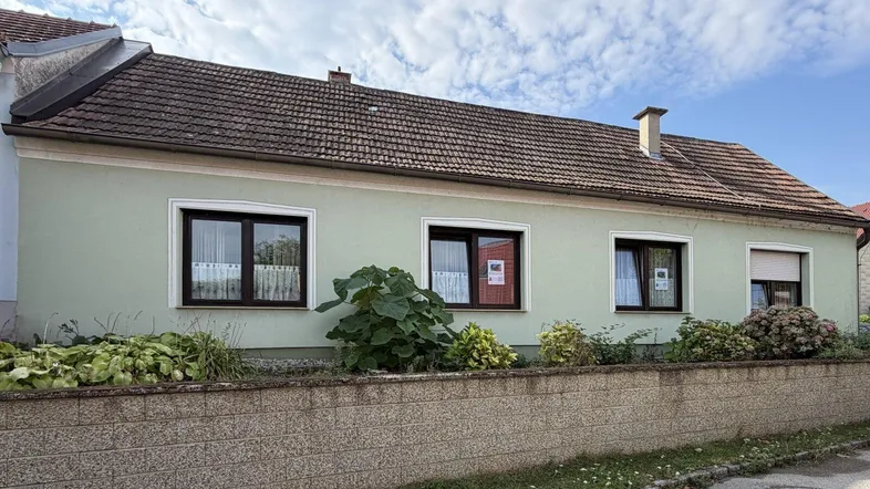 Traditionelles Einfamilienhaus mit hellgrüner Fassade und braunem Ziegeldach unter blauem Himmel.
