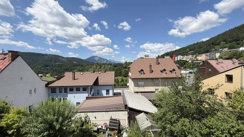 Weitblick über Dächer, grüne Hügel und Berge unter blauem Himmel mit Wolken.