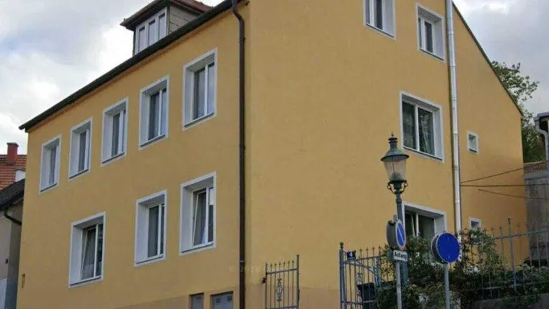 Gelbes Mehrfamilienhaus mit mehreren Fenstern an einer leicht ansteigenden Straße unter bewölktem Himmel.