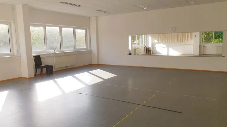 Großer, heller Mehrzweckraum mit vielen Fenstern, Spiegelfläche und Vinylboden, ideal für verschiedene Aktivitäten.