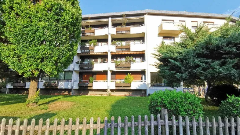 Mehrfamilienhaus mit mehreren Balkonen und gepflegter Grünfläche, umgeben von Bäumen und einem Holzzaun.