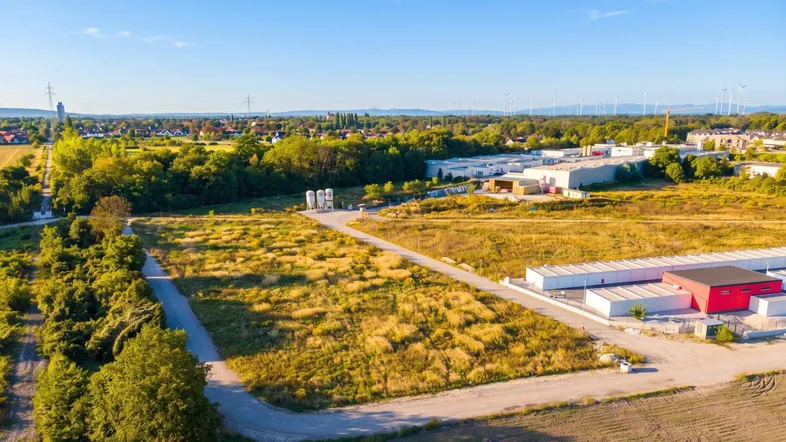 Luftaufnahme eines großen unbebauten Grundstücks mit umliegenden Feldern und Industriegebäuden im Hintergrund.