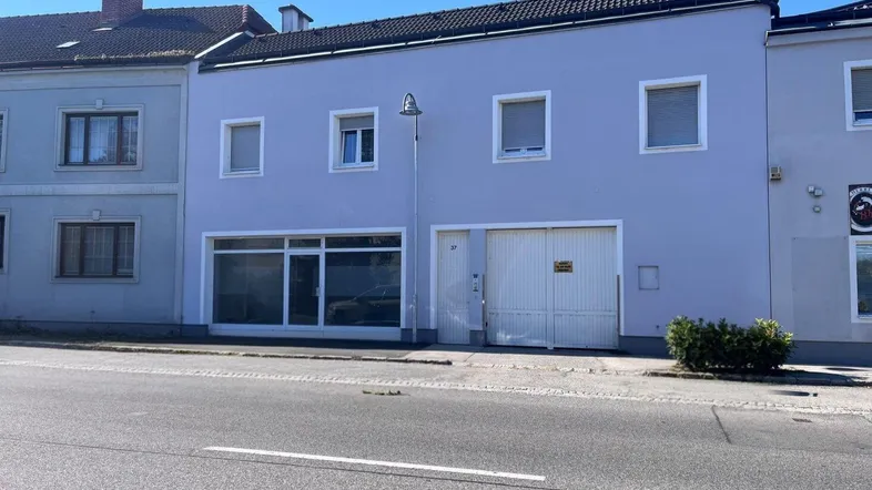Gebäudeansicht mit Ladenfront und Garagentor an einer Straße, in einem hellen Grauton gestrichen.